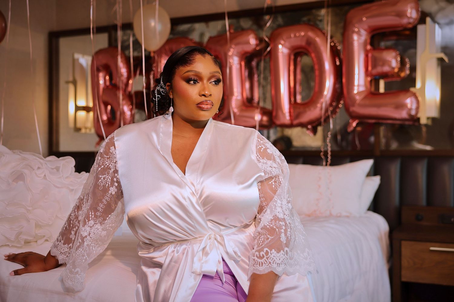 Woman in white satin bridal robe sits before rose gold BRIDE balloon letters on a decorated bed.