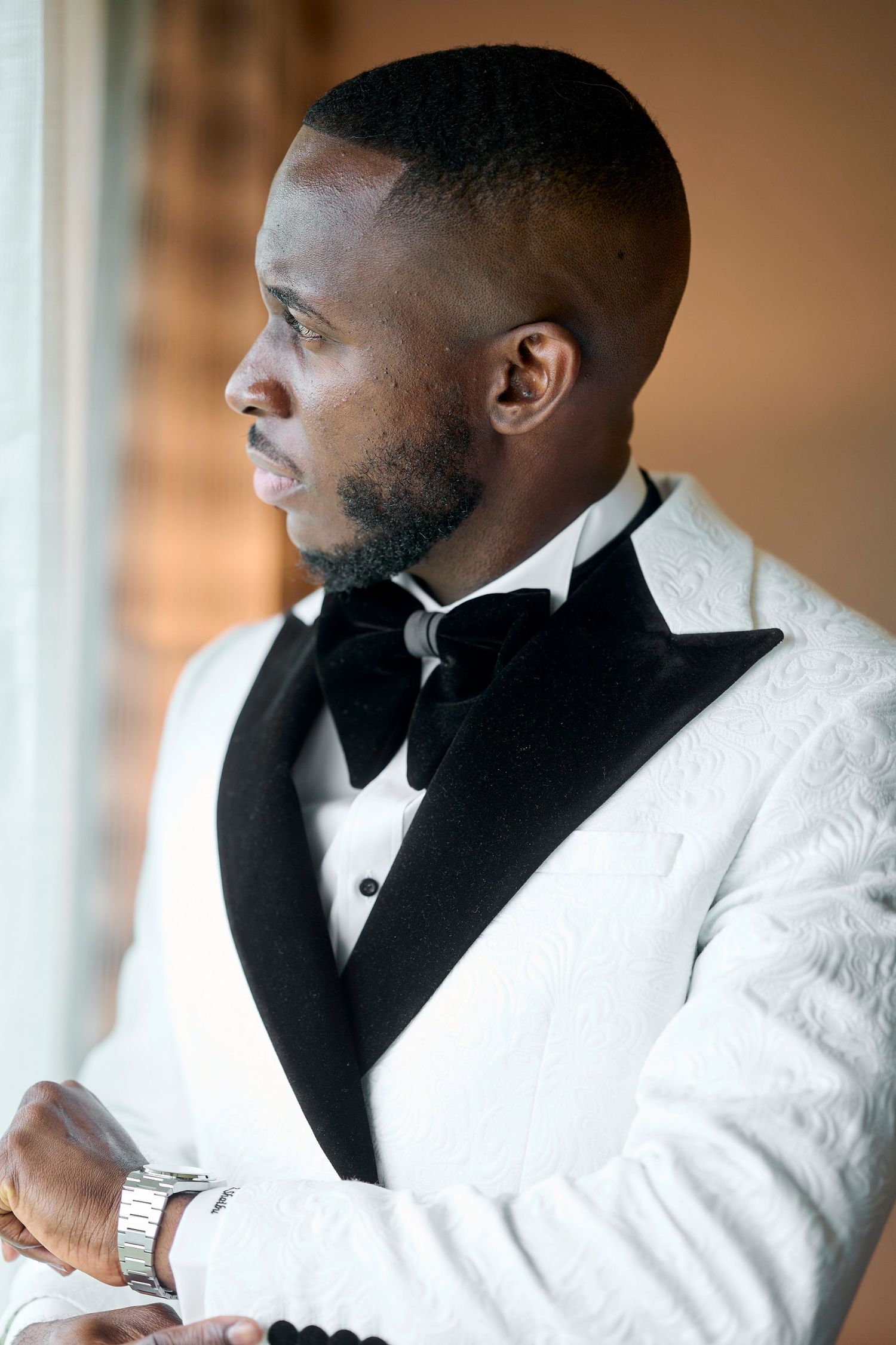 Elegant groom in white floral tuxedo jacket with black lapels and bow tie gazing thoughtfully out a window.