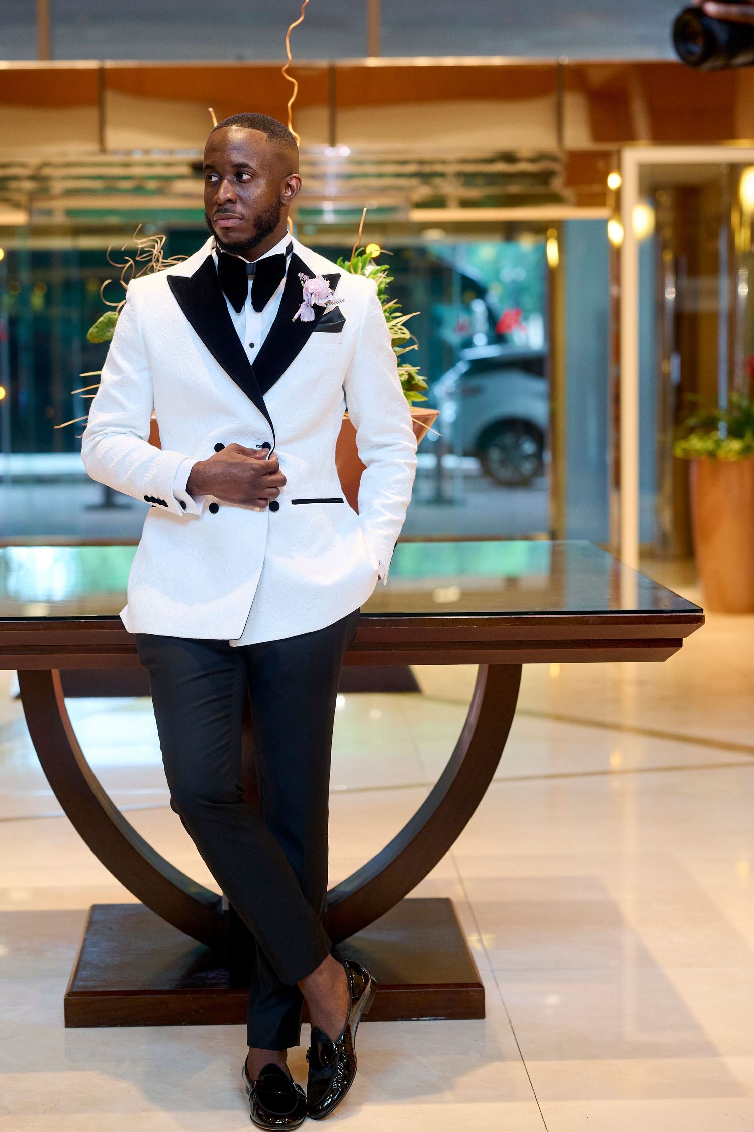 Elegant Black man in white tuxedo jacket with black lapels and bow tie leans against a wooden table in a hotel lobby.