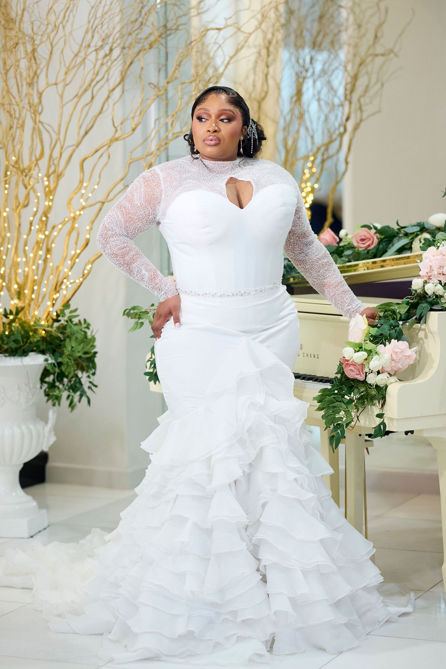Plus-size bride in white mermaid wedding gown with lace sleeves and ruffled skirt posing elegantly.