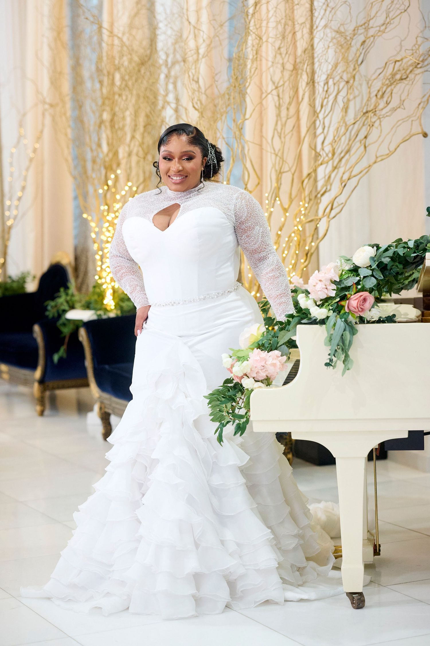 Bride in white mermaid gown with lace sleeves and ruffled skirt, holding bouquet beside a white piano.