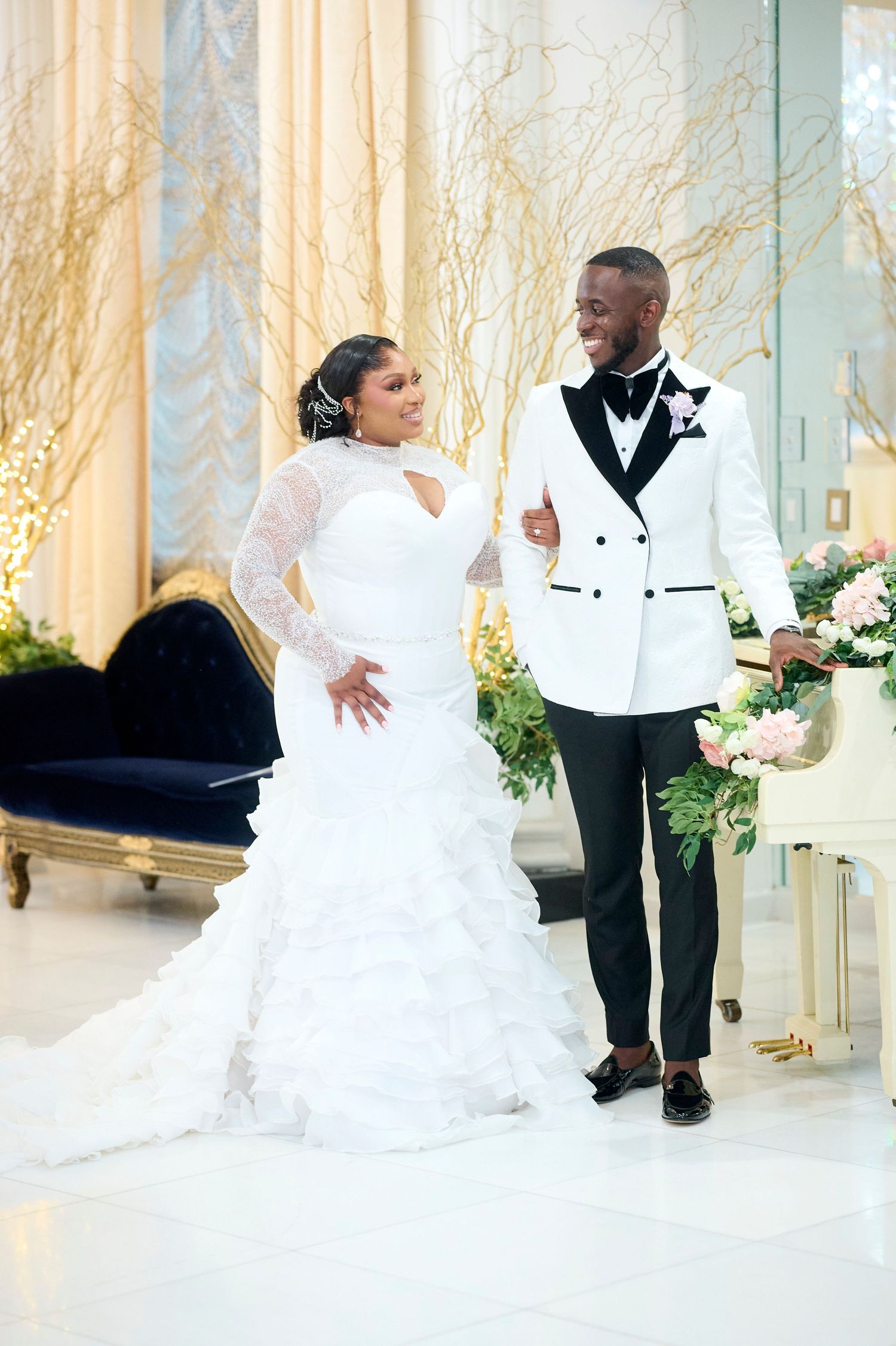 Elegant Black couple in wedding attire, bride in white ruffled gown and groom in white tuxedo jacket, posing in ornate venue.