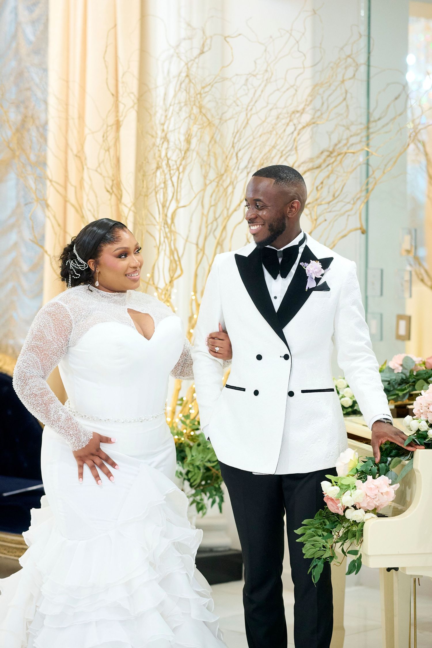 A bride in a lace mermaid gown and groom in a white tuxedo jacket smile together at their elegant wedding ceremony.