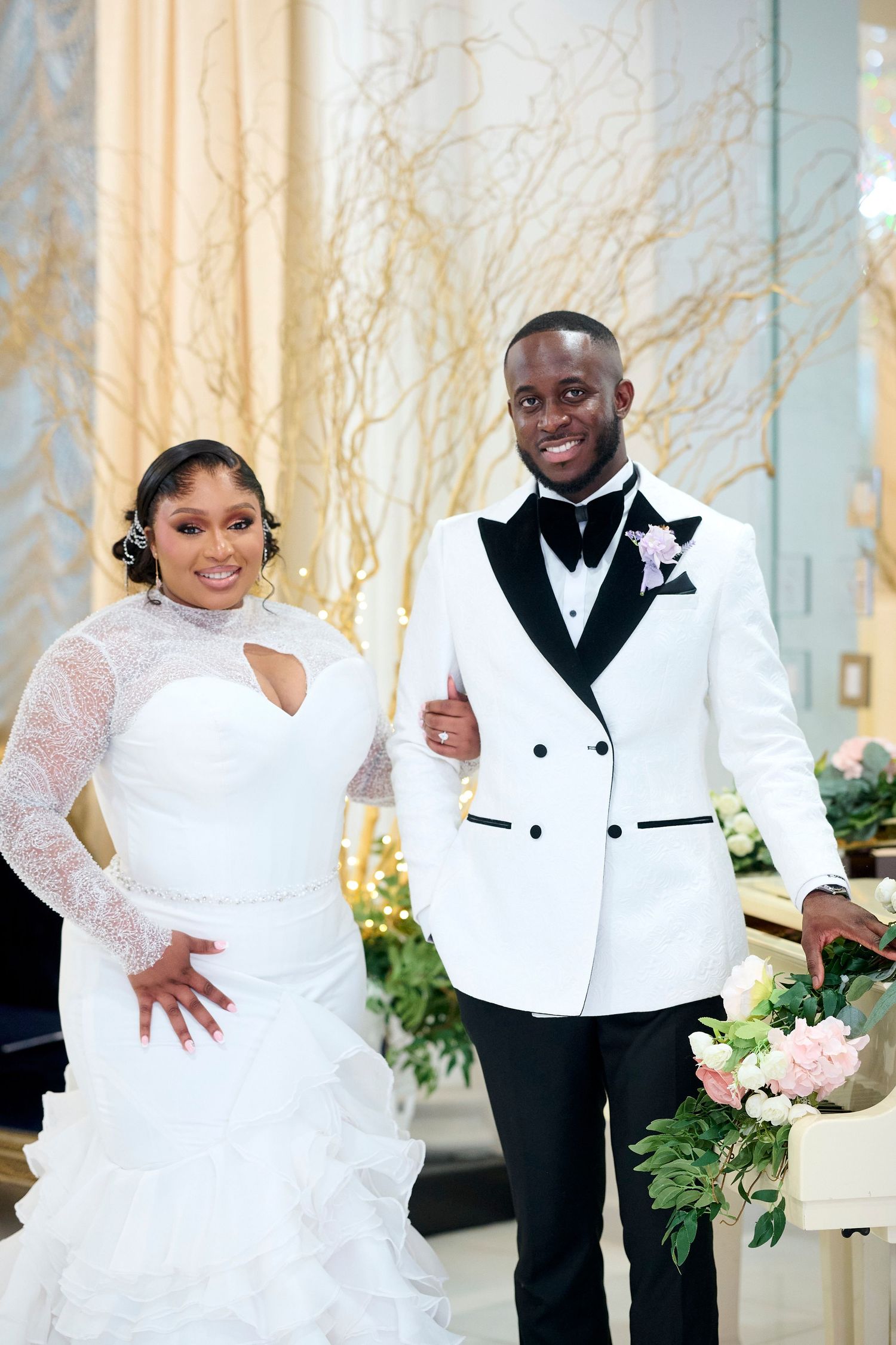 Smiling Black bride and groom posing at altar, she in ruffled white gown, he in white double-breasted tuxedo jacket.