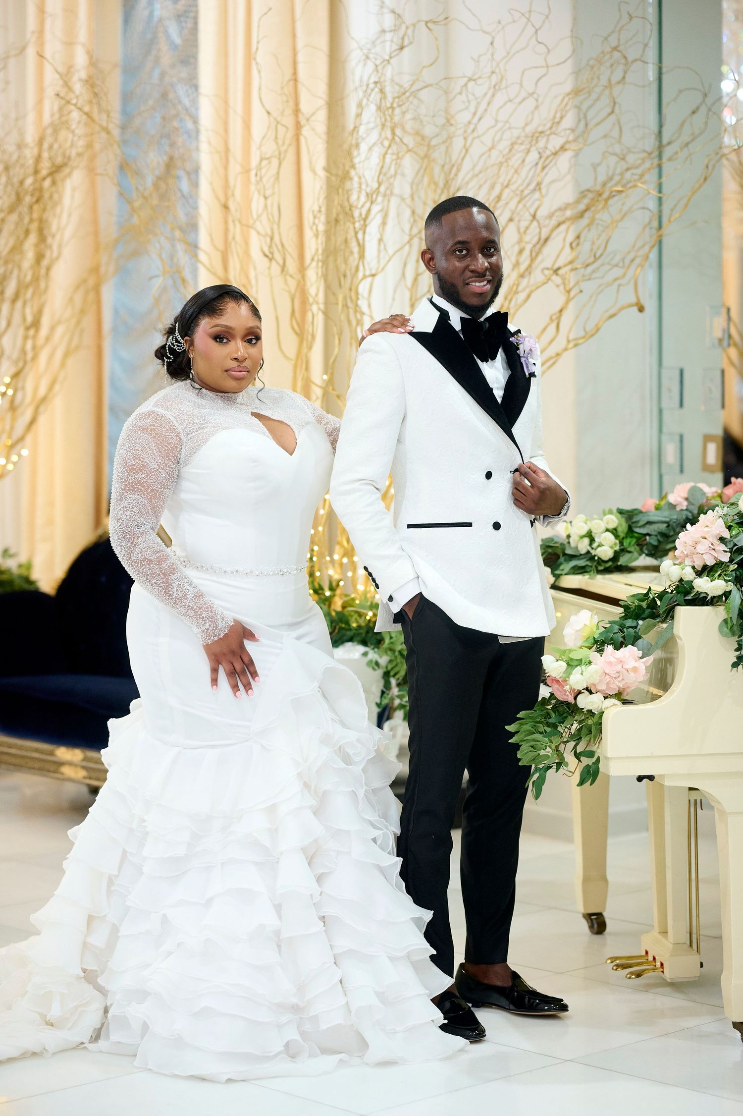 Elegant Black couple posing at wedding, bride in ruffled lace gown, groom in white tuxedo jacket with black lapels.