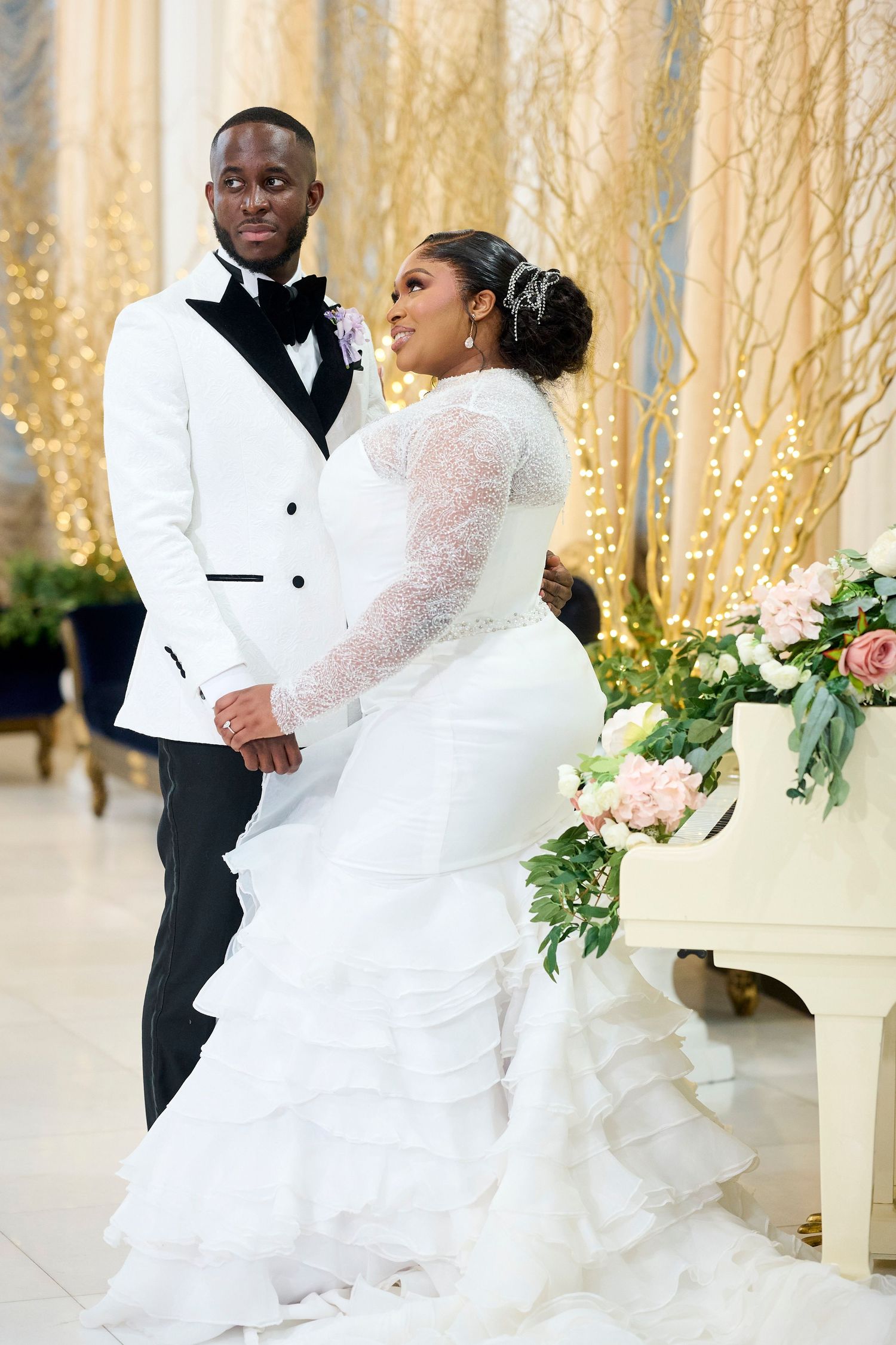 A bride and groom pose elegantly in wedding attire, she in a white mermaid gown, he in a white tuxedo, by a floral piano.
