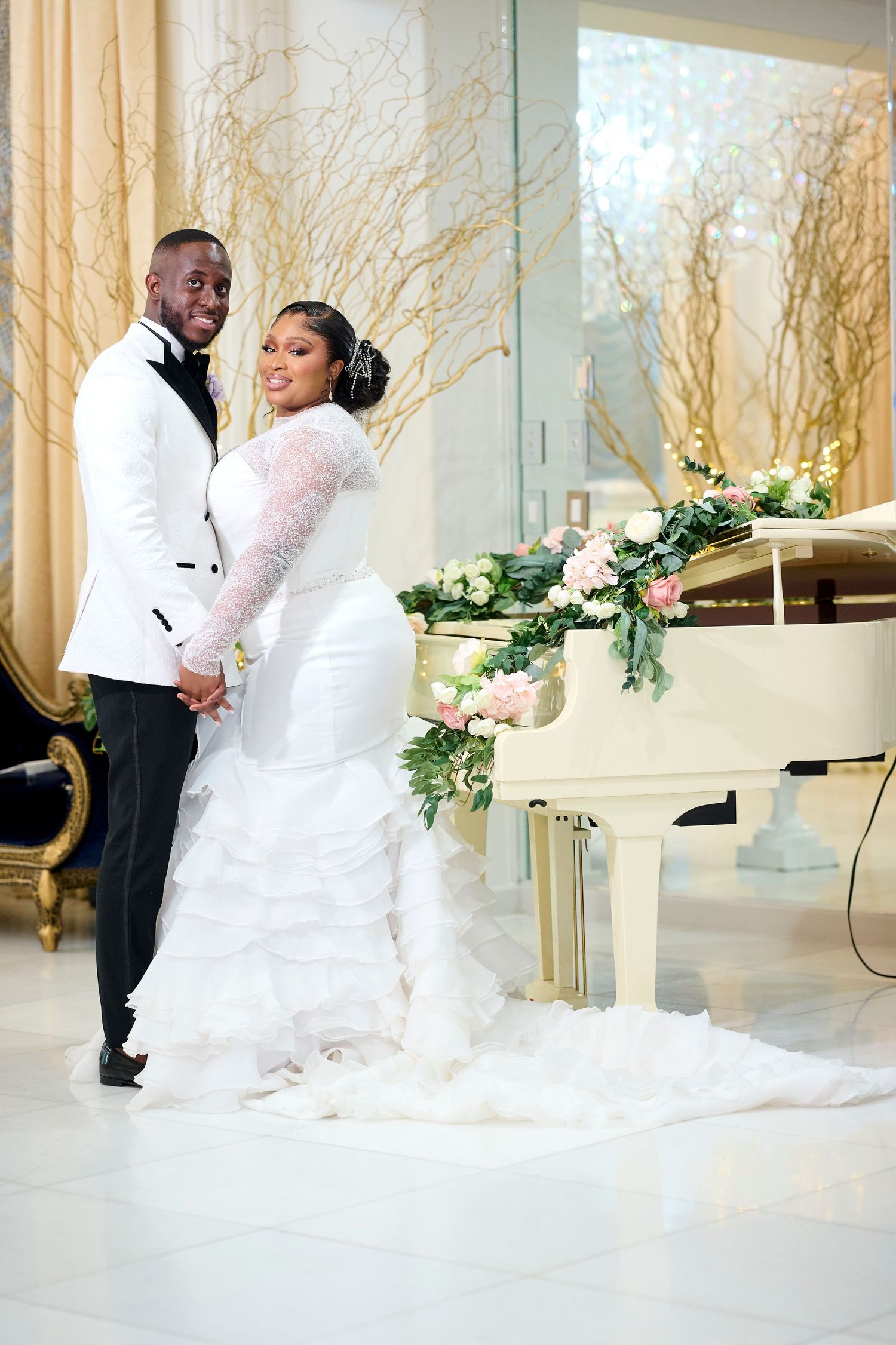 Elegant wedding couple posing by a white grand piano adorned with flowers in a luxurious gold and white venue.