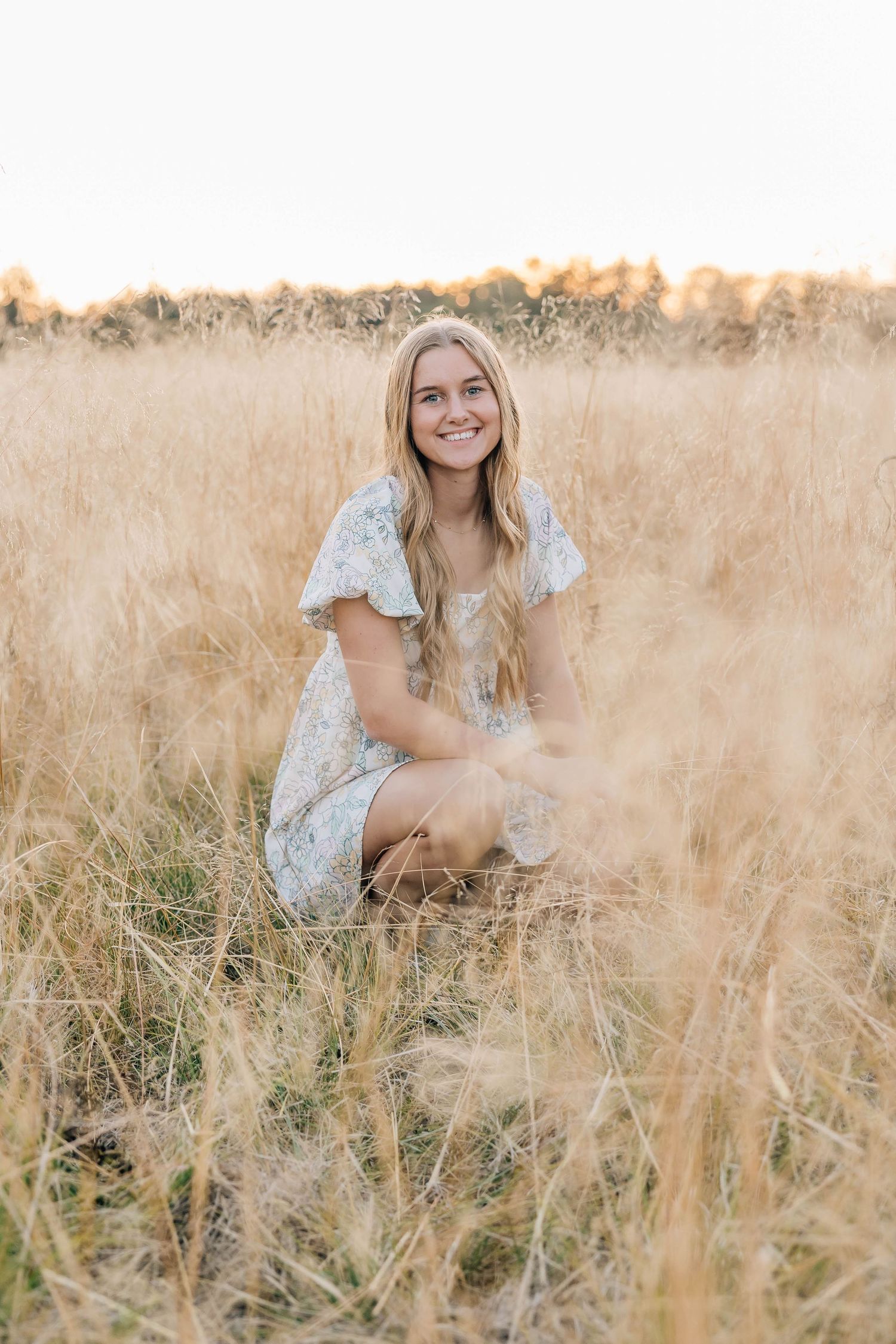 Smiling blonde teenage girl crouching in tall golden grass field wearing floral summer dress.