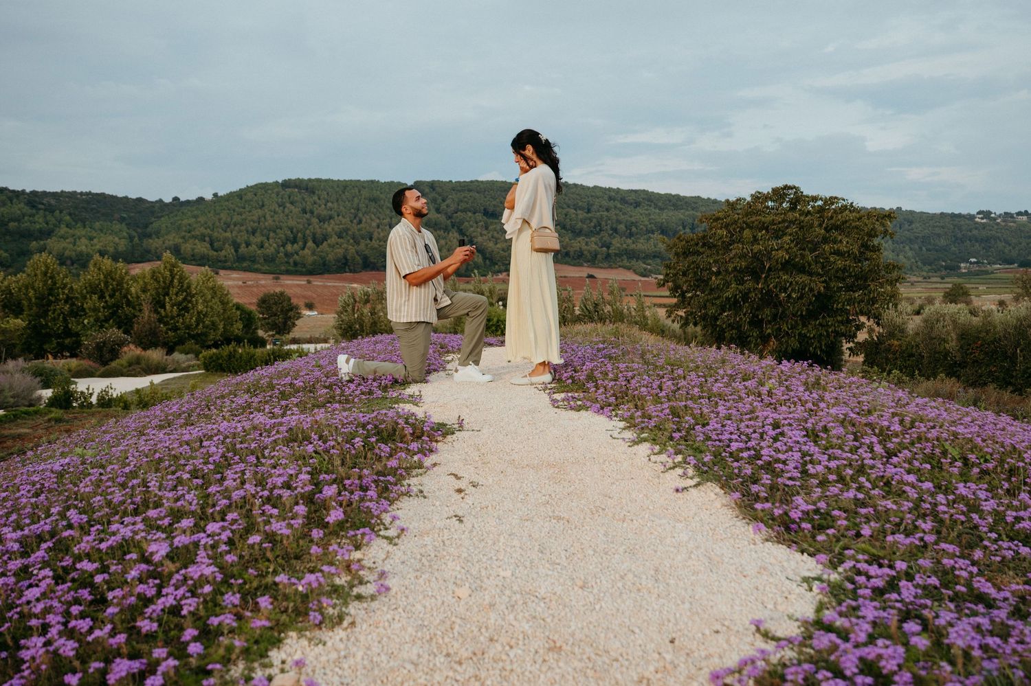 Couple sharing a romantic moment on a gravel path surrounded by purple wildflowers with rolling hills backdrop.