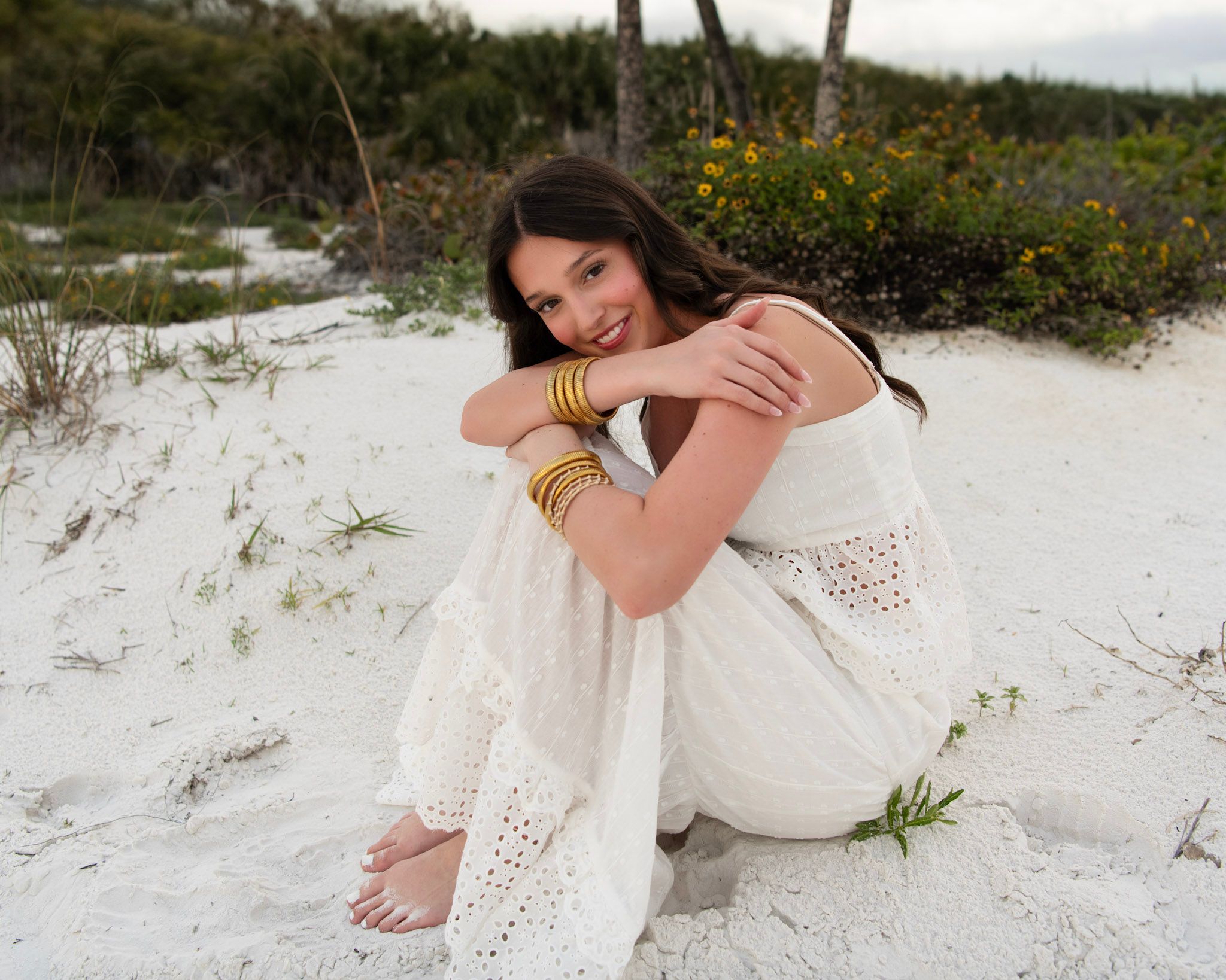 Young woman in white eyelet dress sitting on white sand beach, wearing gold bangles, smiling outdoors.