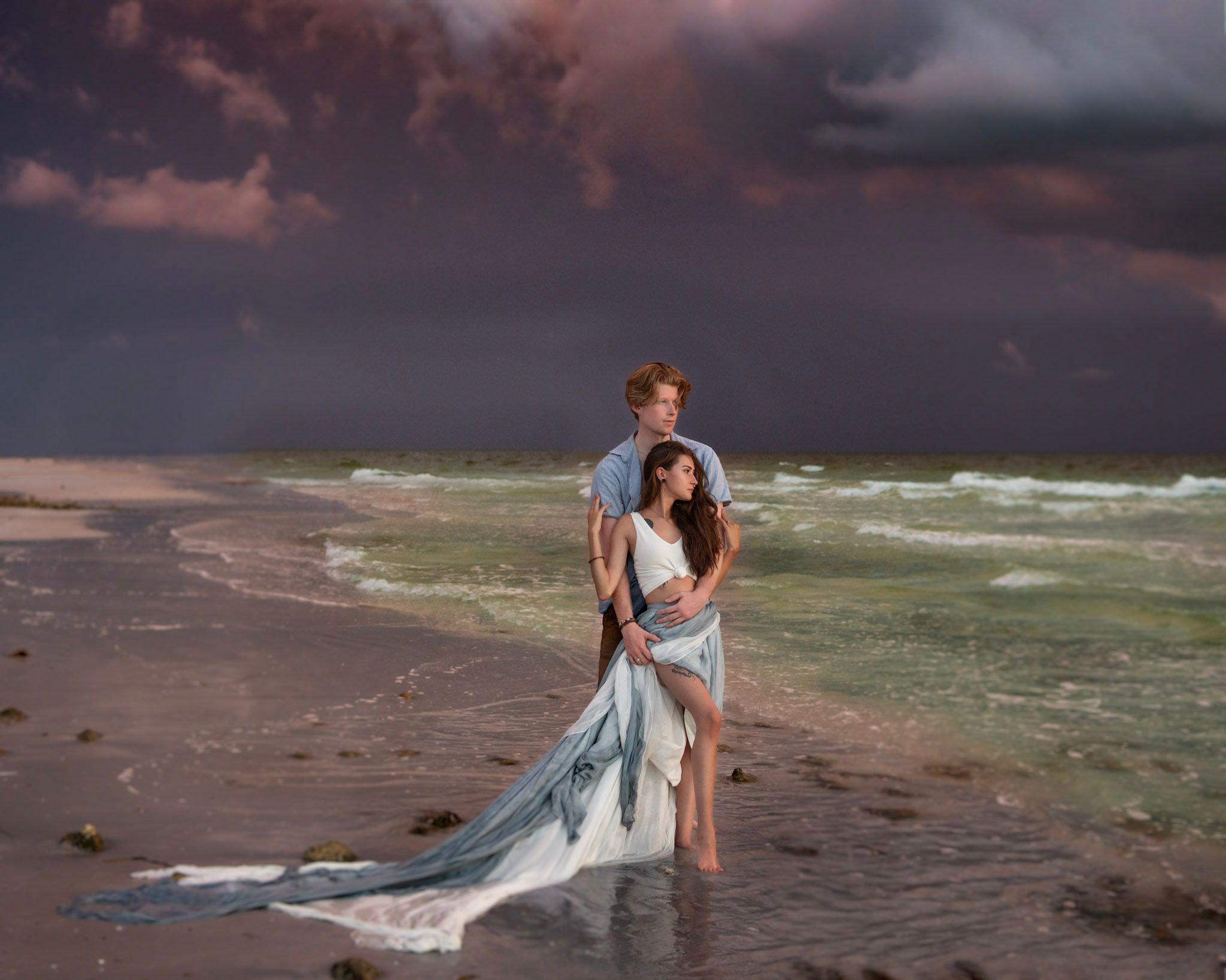 Romantic couple embracing on a stormy beach at sunset, woman in flowing white gown with dramatic clouds overhead.