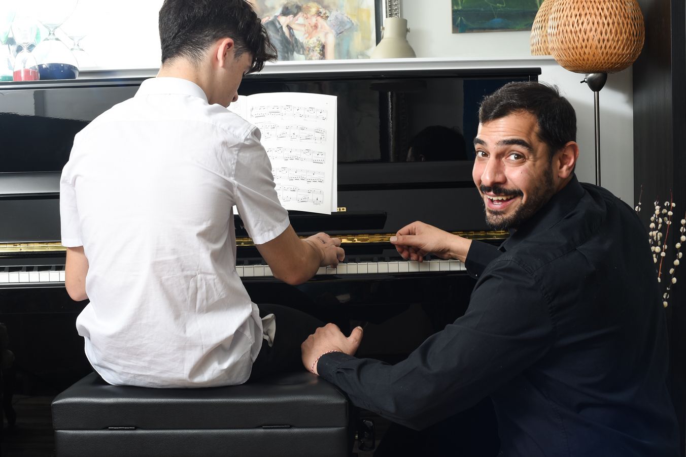 A smiling man assists a young pianist at a black piano with sheet music. The room is cozy, with warm lighting and decorative elements.