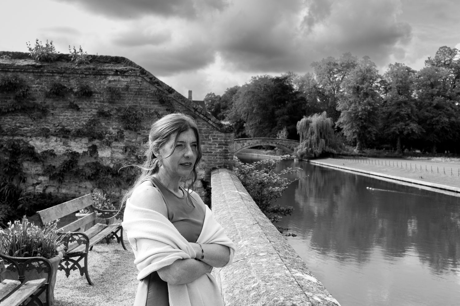 Black and white photo of a woman standing by a serene canal with historic stone walls and lush trees in background.