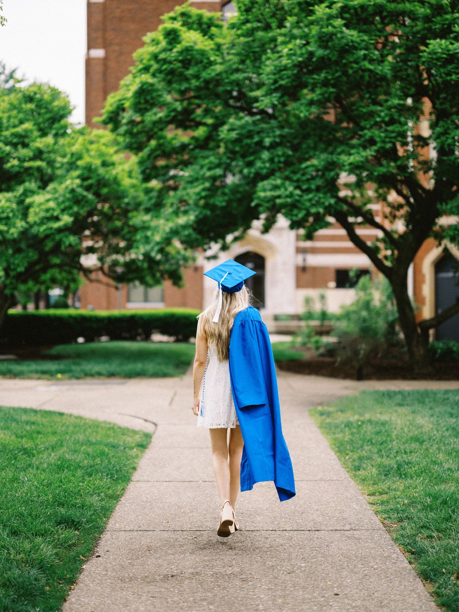 A graduate in blue cap and gown walks along a campus pathway surrounded by lush green trees and brick buildings.