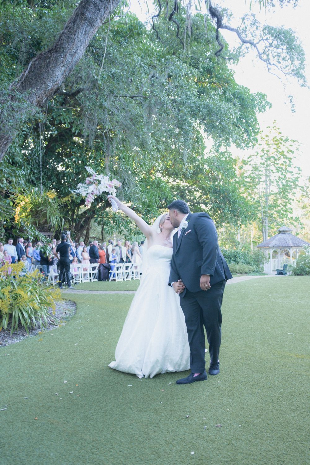 Bride raises bouquet while groom kisses her cheek on lush green lawn with guests seated under large oak trees.