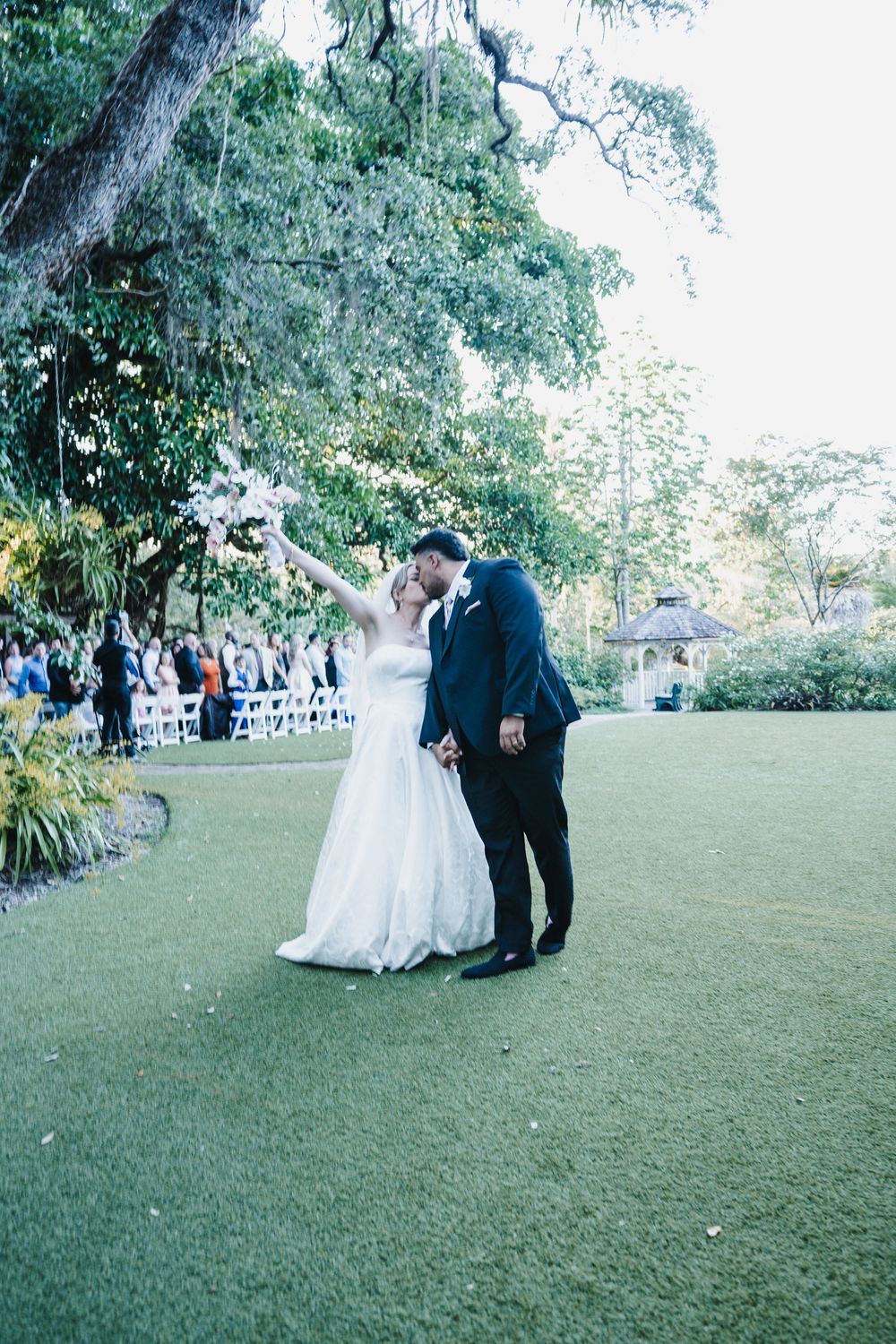 Bride raises bouquet while groom gazes at her on lush garden lawn after outdoor wedding ceremony.