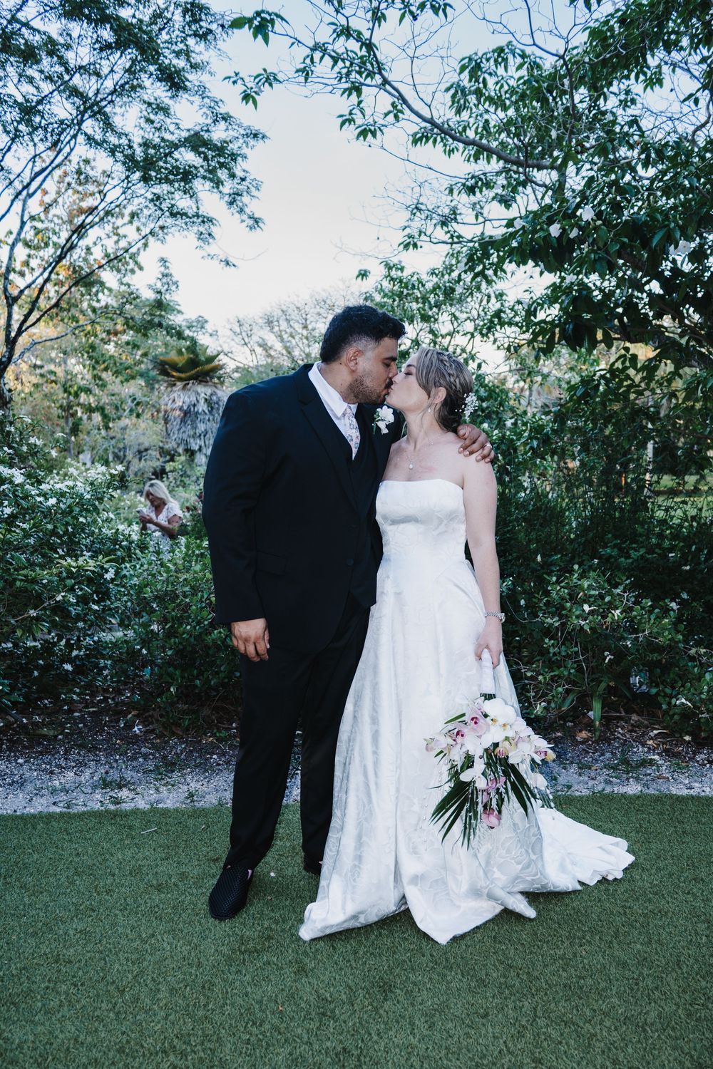 A bride and groom share a kiss outdoors, surrounded by lush greenery, as she holds a beautiful floral bouquet.