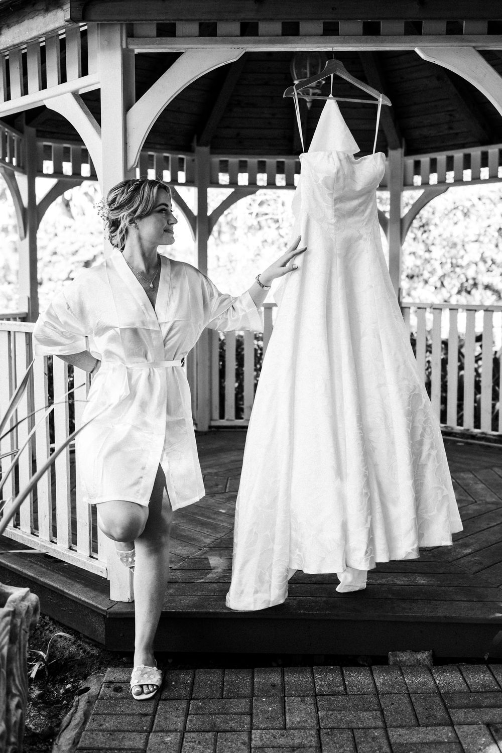Bride in white robe admiring her wedding dress hanging in a gazebo, black and white photo.