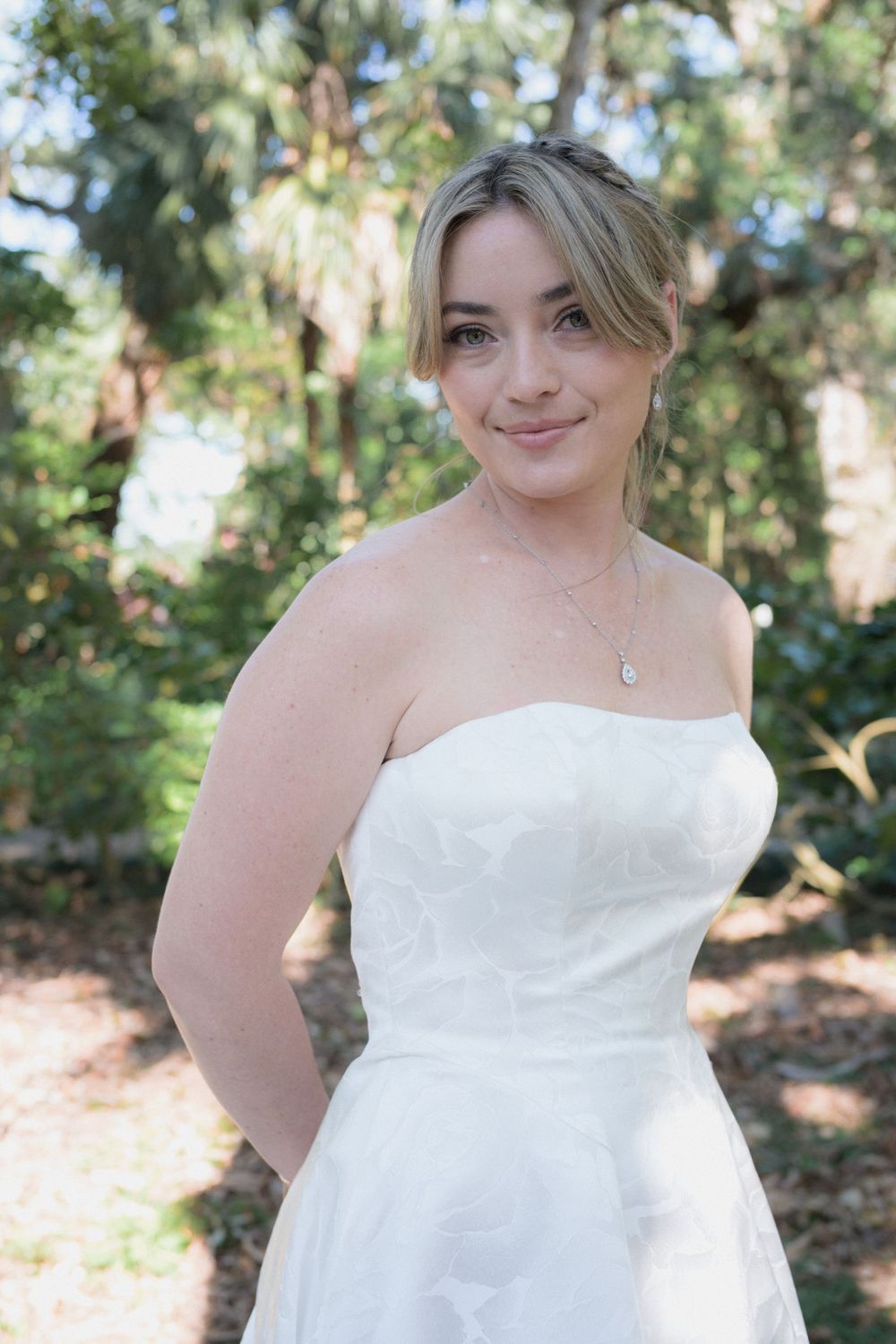 Smiling bride in strapless white gown with updo hairstyle and delicate necklace posing outdoors among lush greenery.