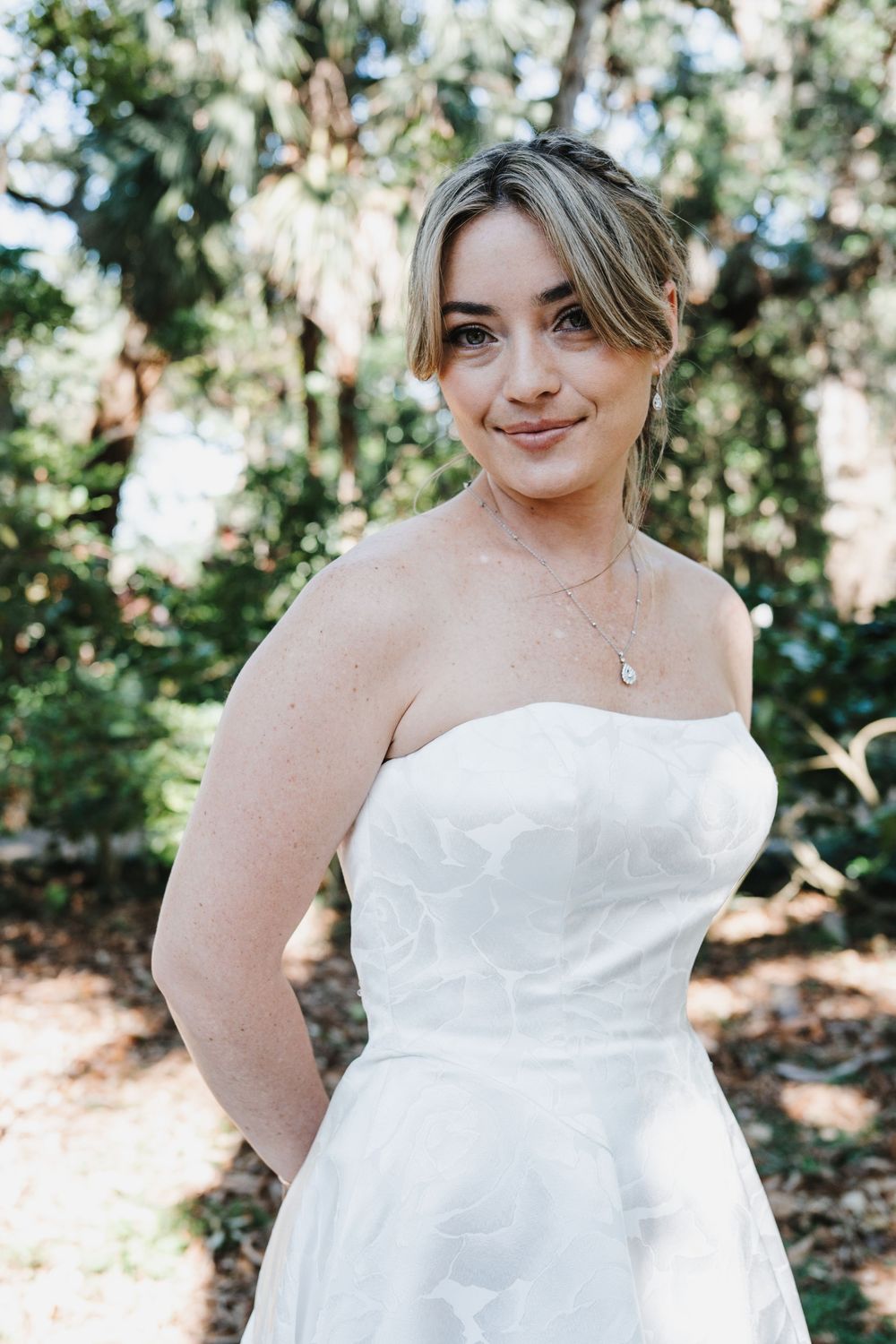 A smiling bride in a strapless white gown poses outdoors among lush green trees on her wedding day.
