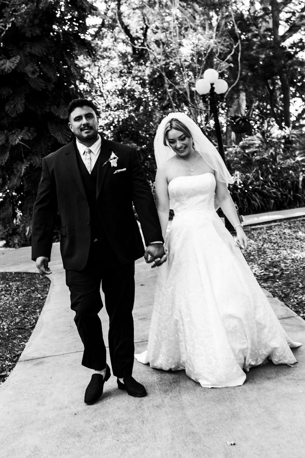 Black and white photo of a smiling bride and groom holding hands walking down a path outdoors on their wedding day.