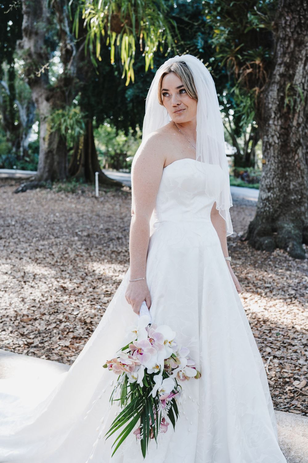 Bride in strapless white gown and veil holds floral bouquet outdoors among trees.