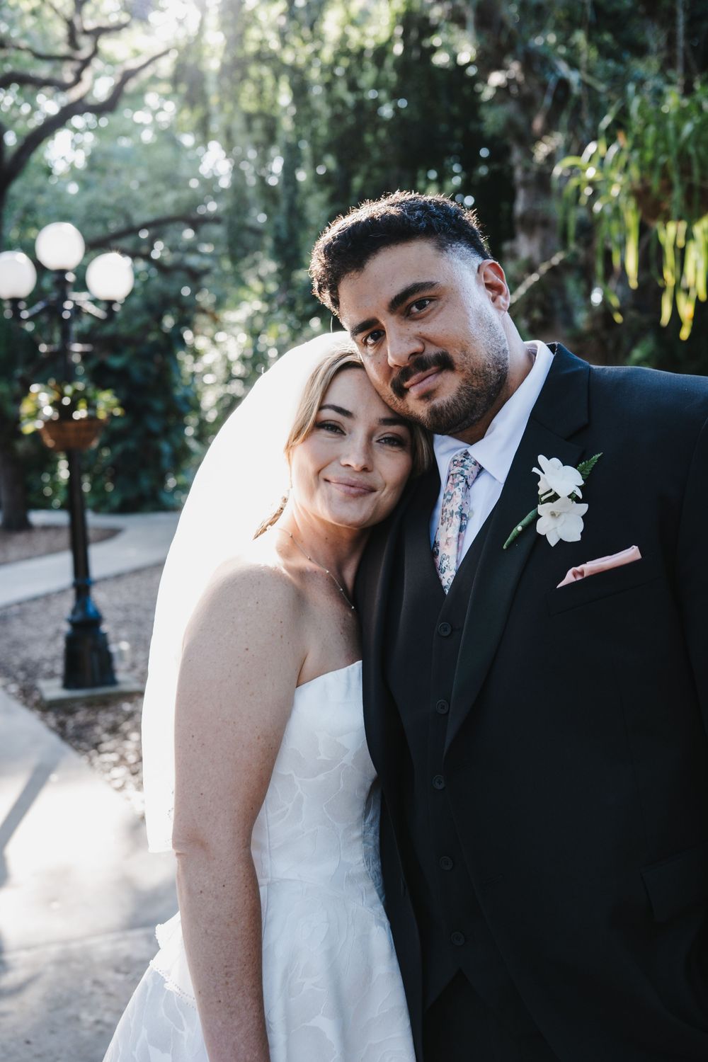 Smiling bride and groom pose together outdoors in wedding attire among lush trees.