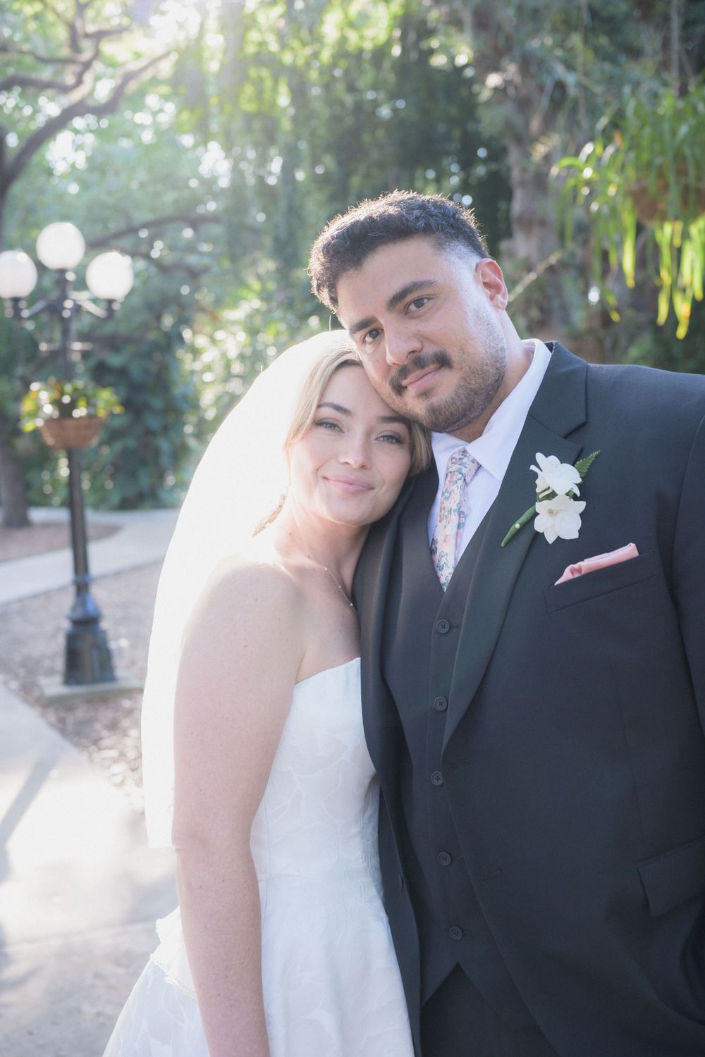 Bride and groom posing together outdoors, groom in dark suit with white boutonniere.
