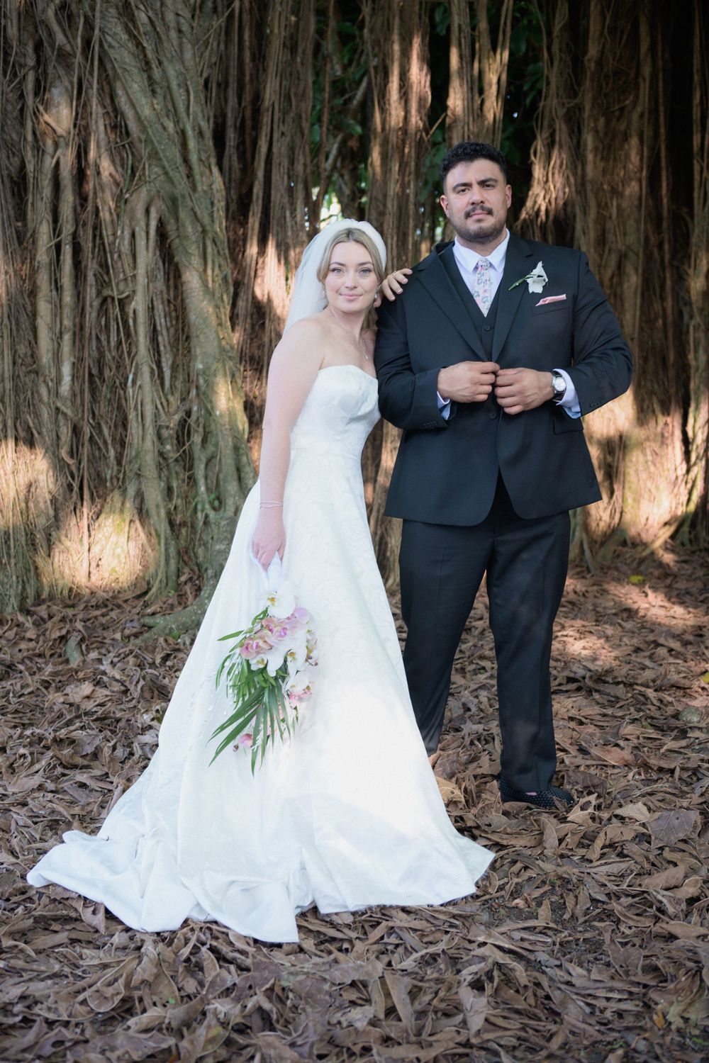 Bride and groom posing together in wooded setting, she in white strapless gown holding bouquet, he in dark suit.