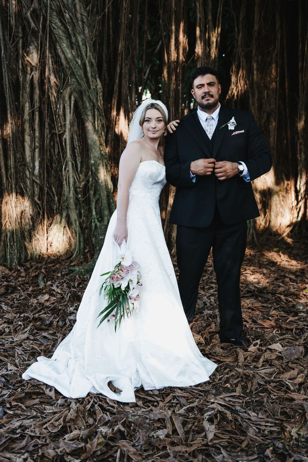 Bride and groom pose together in white gown and dark suit among tall trees with fallen leaves.