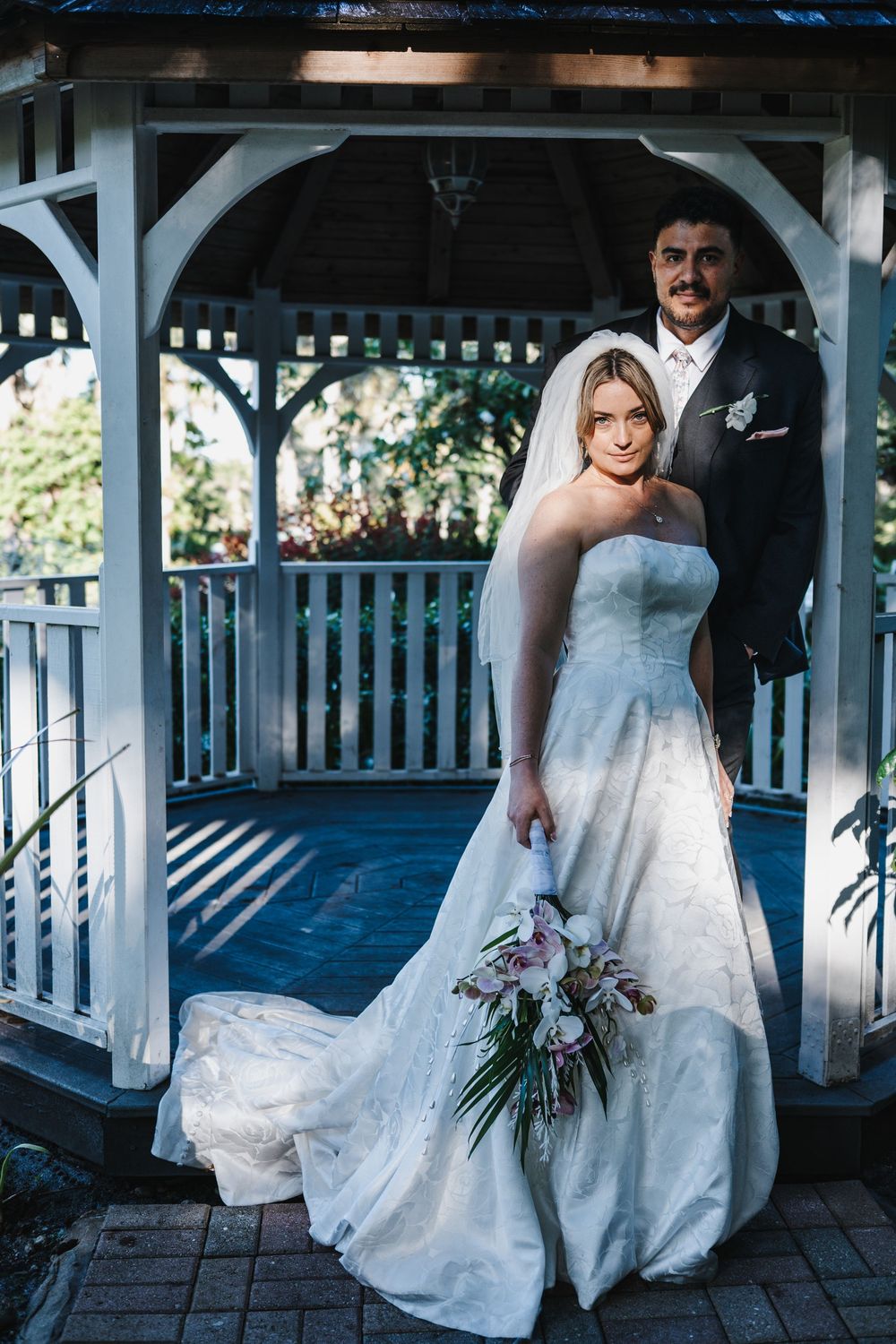 Bride in strapless white gown holding bouquet stands with groom in dark suit inside elegant white gazebo.