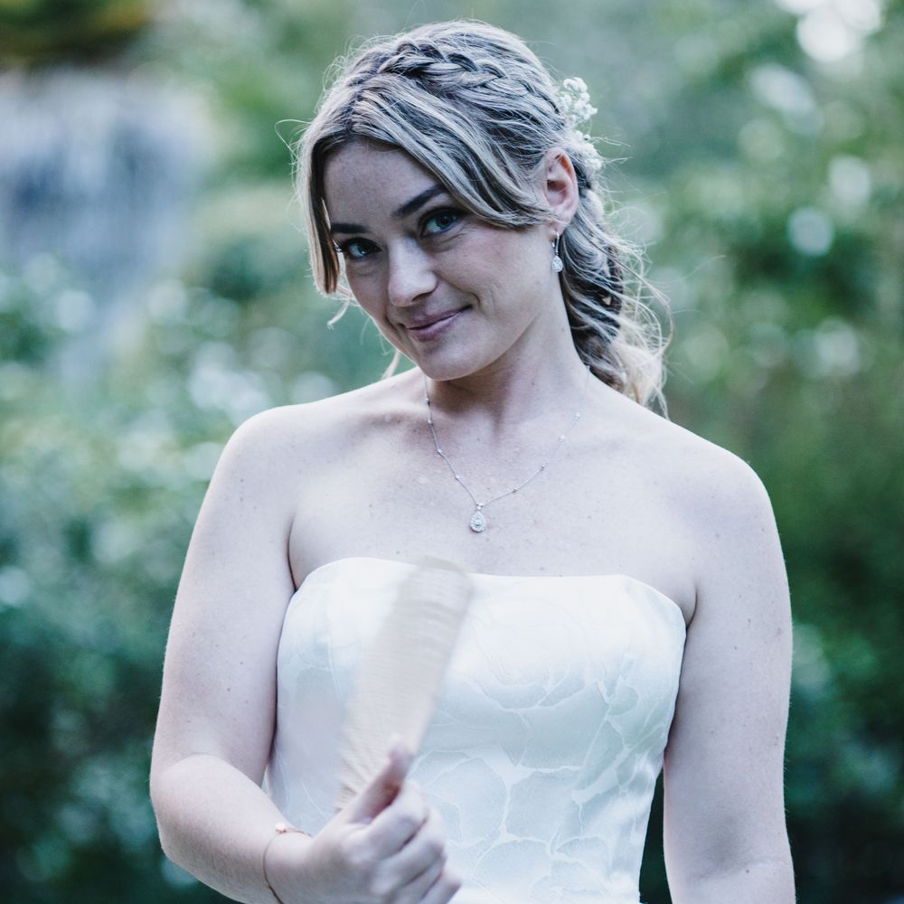 A young woman in a strapless white bridal gown with braided updo hairstyle smiling outdoors.