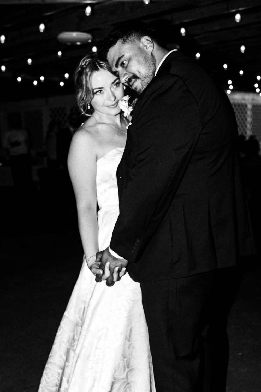 Black and white photo of a bride and groom holding hands and dancing at their wedding reception under string lights.