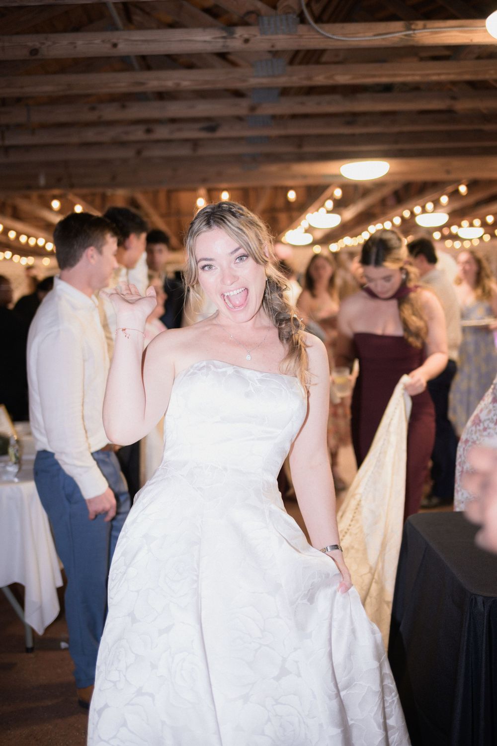 Bride in strapless white gown dancing joyfully at rustic barn wedding reception with string lights.