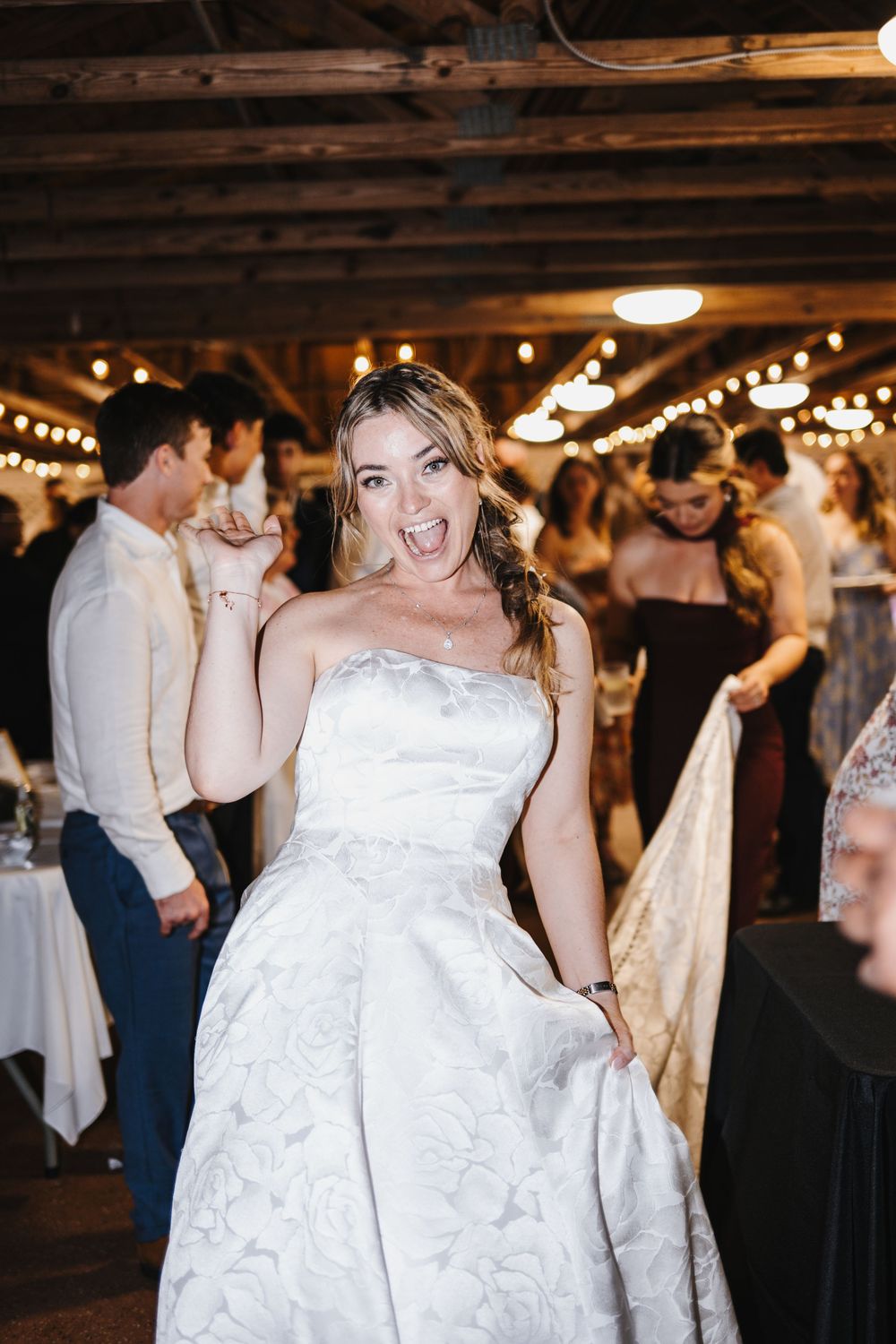 Joyful bride in a strapless floral white gown dancing at a rustic wedding reception with string lights.