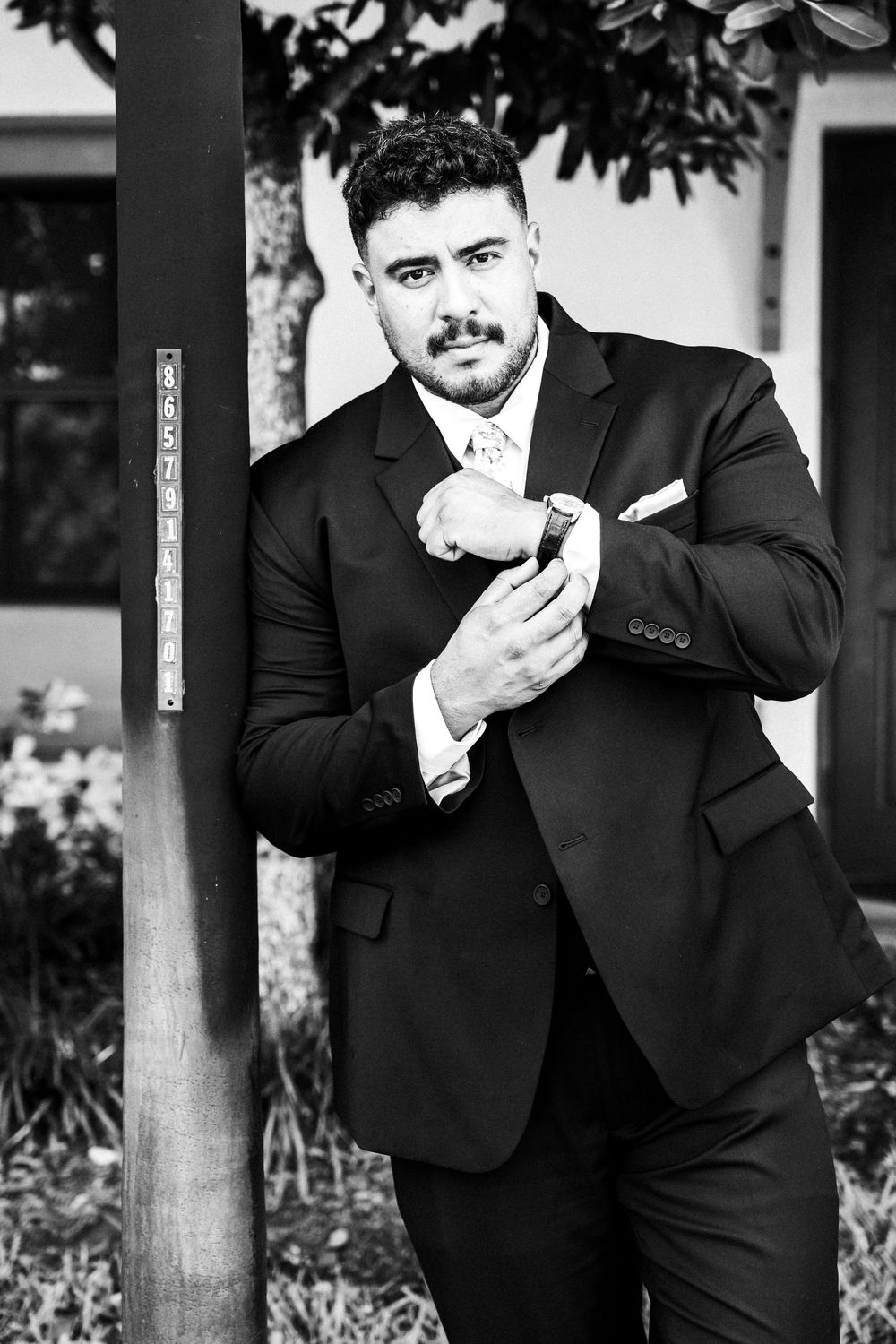 A well-dressed man in a black suit adjusts his cufflinks while leaning against a wooden post outdoors.