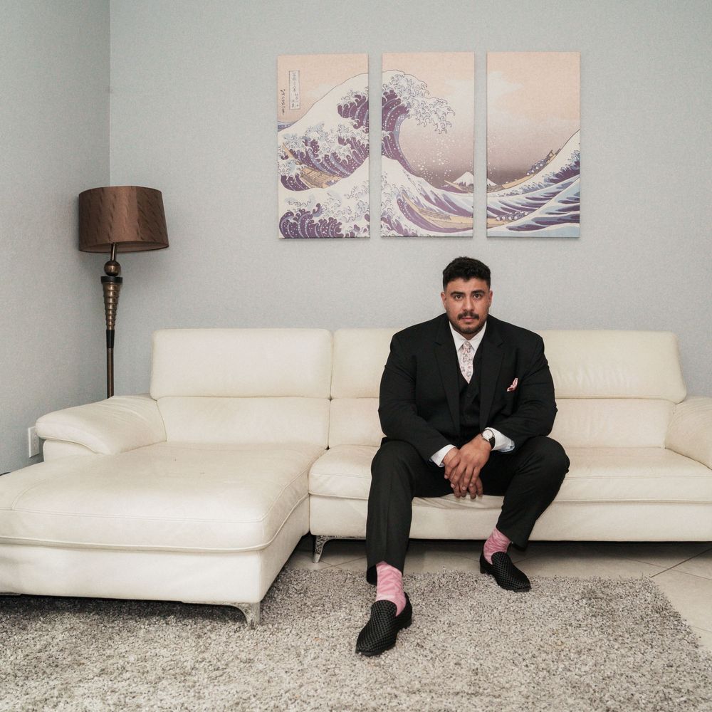 Man in black suit with pink socks sits on white sectional sofa beneath The Great Wave art triptych.