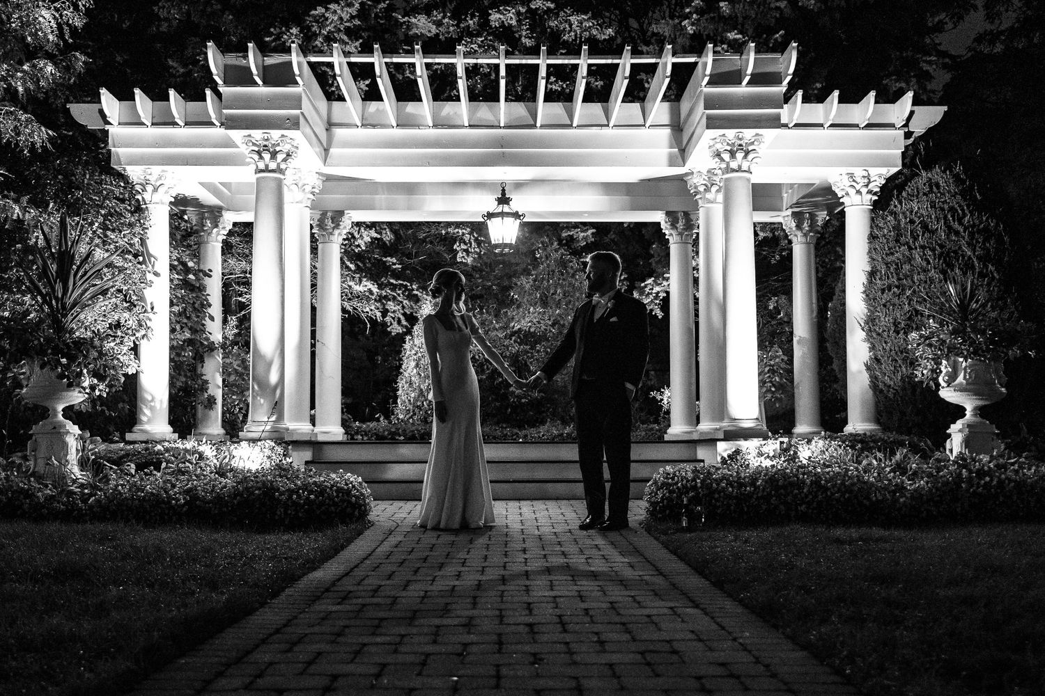 Black and white photo of a couple holding hands under an illuminated white pergola at night.