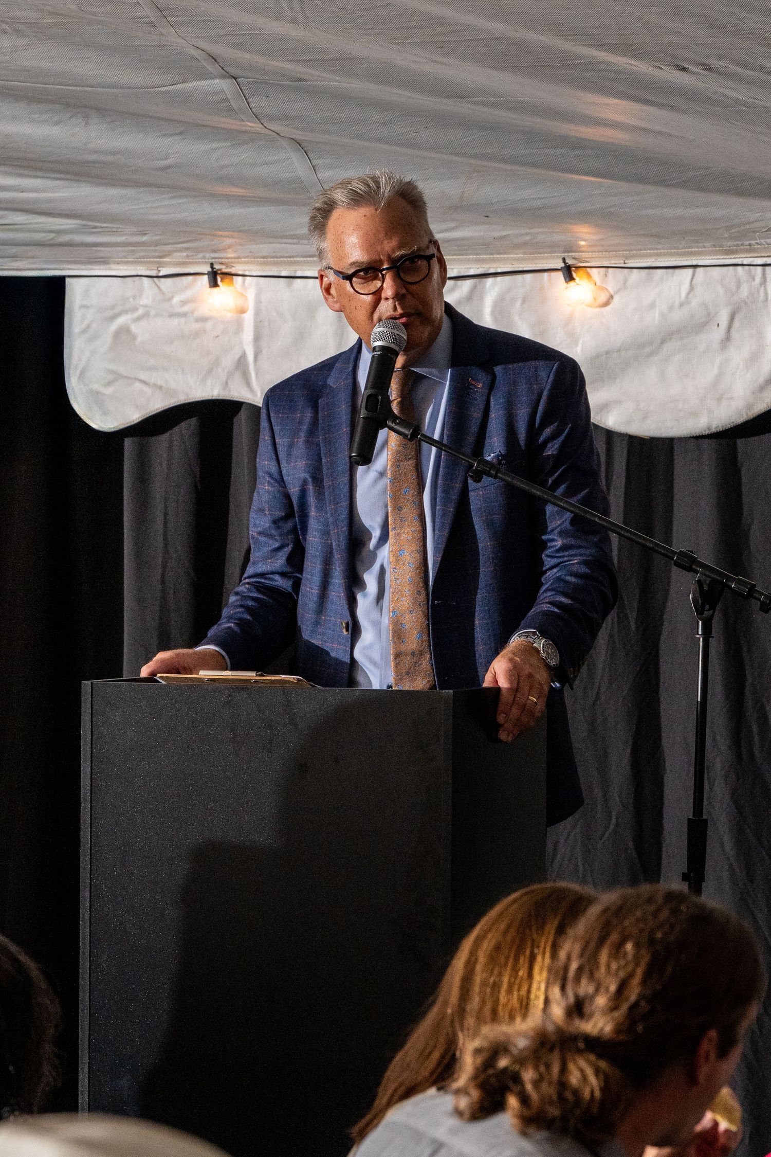 Man in blue suit and glasses speaks at podium with microphone at formal indoor event under tent.