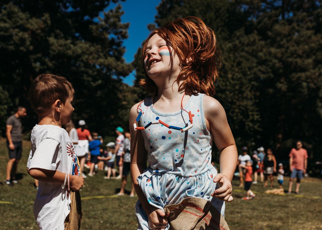 A joyful red-haired girl laughs and dances outdoors at a sunny park event with children playing in the background.