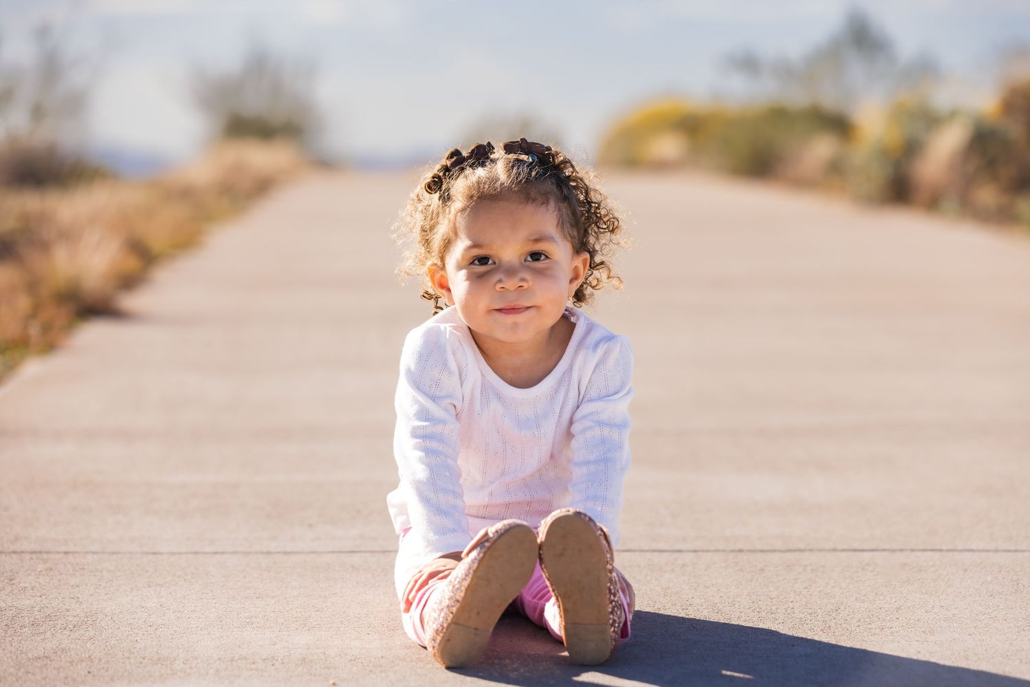 A cheerful toddler girl in pink pants and white top walks alone on a sunny desert road surrounded by dry brush.