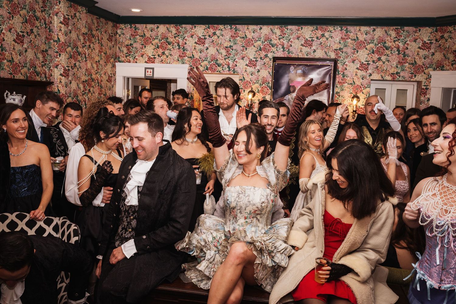 Joyful wedding reception with bride and groom surrounded by celebrating guests in a floral wallpapered room.