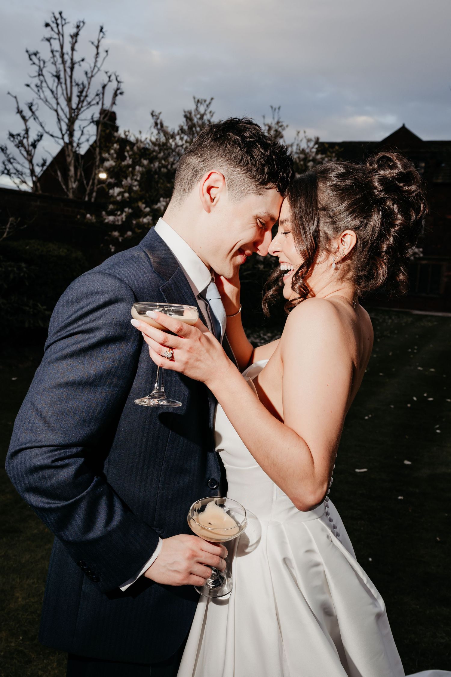 Newlywed couple smiling and touching foreheads while holding champagne glasses outdoors at their wedding reception.