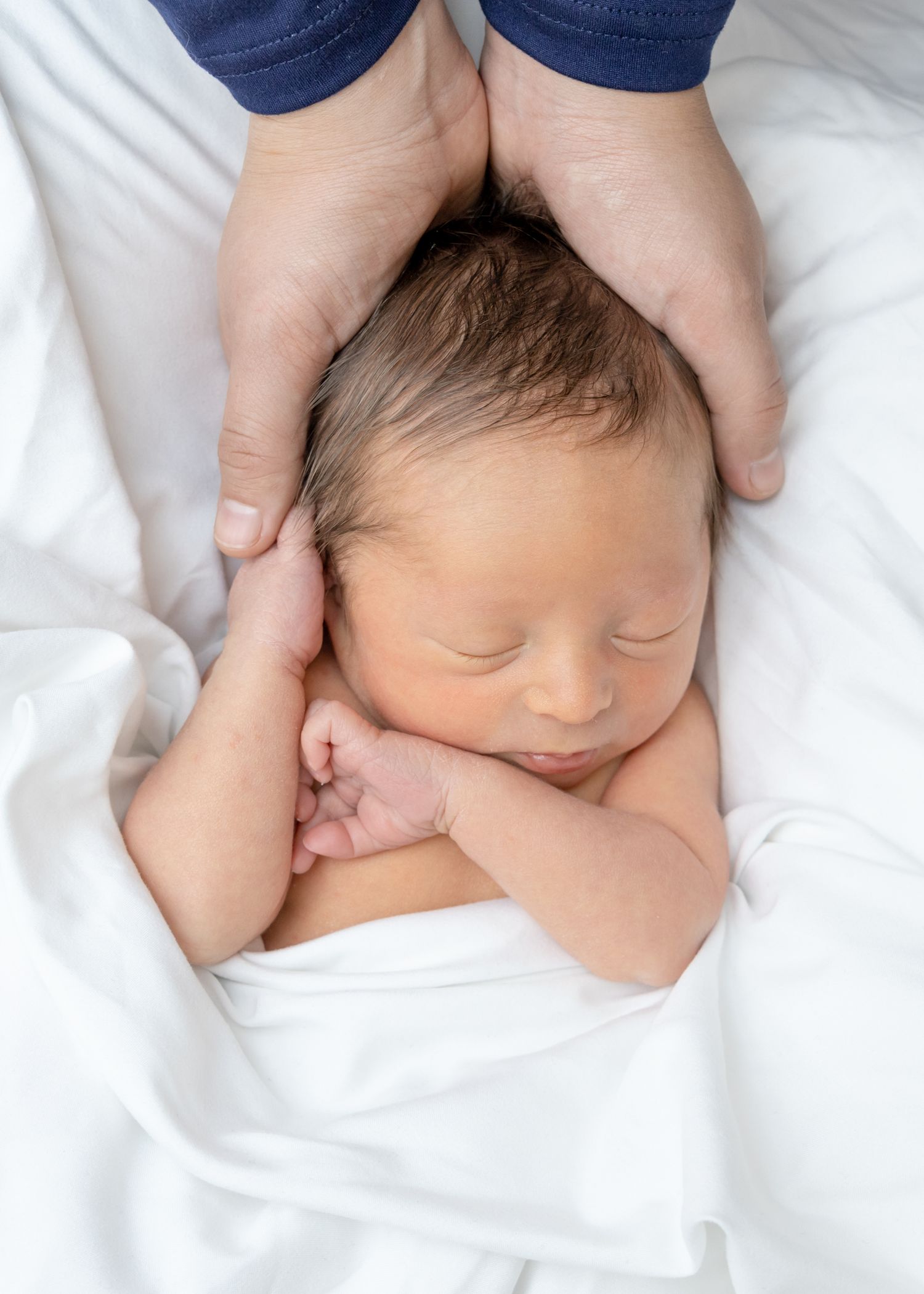A sleeping newborn baby swaddled in white bedding rests peacefully on soft white sheets.
