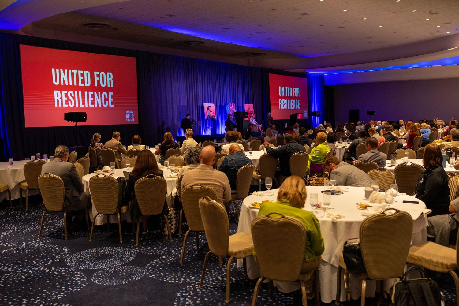 Large conference audience seated at round tables facing a stage with a red screen reading United for Resilience.