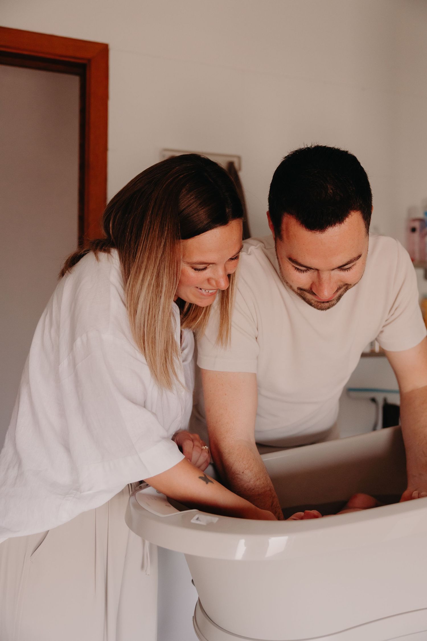 Smiling parents bathing their newborn baby together in a white baby bathtub.