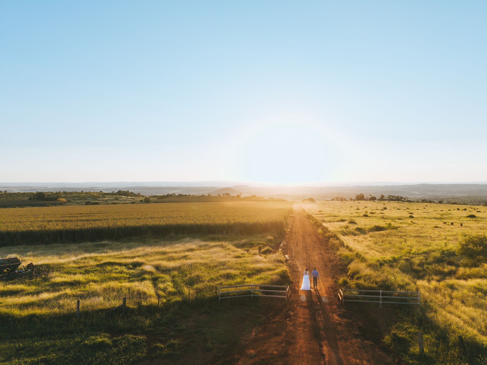 Aerial view of a couple walking along a dirt road through lush green farmland at golden sunset.