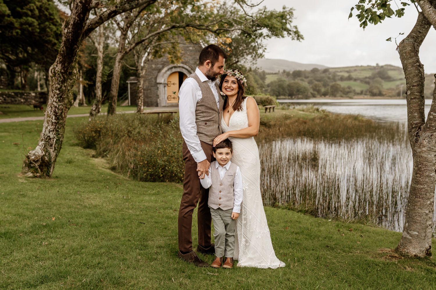 Family wedding portrait by a serene lake with trees and rolling hills in background.