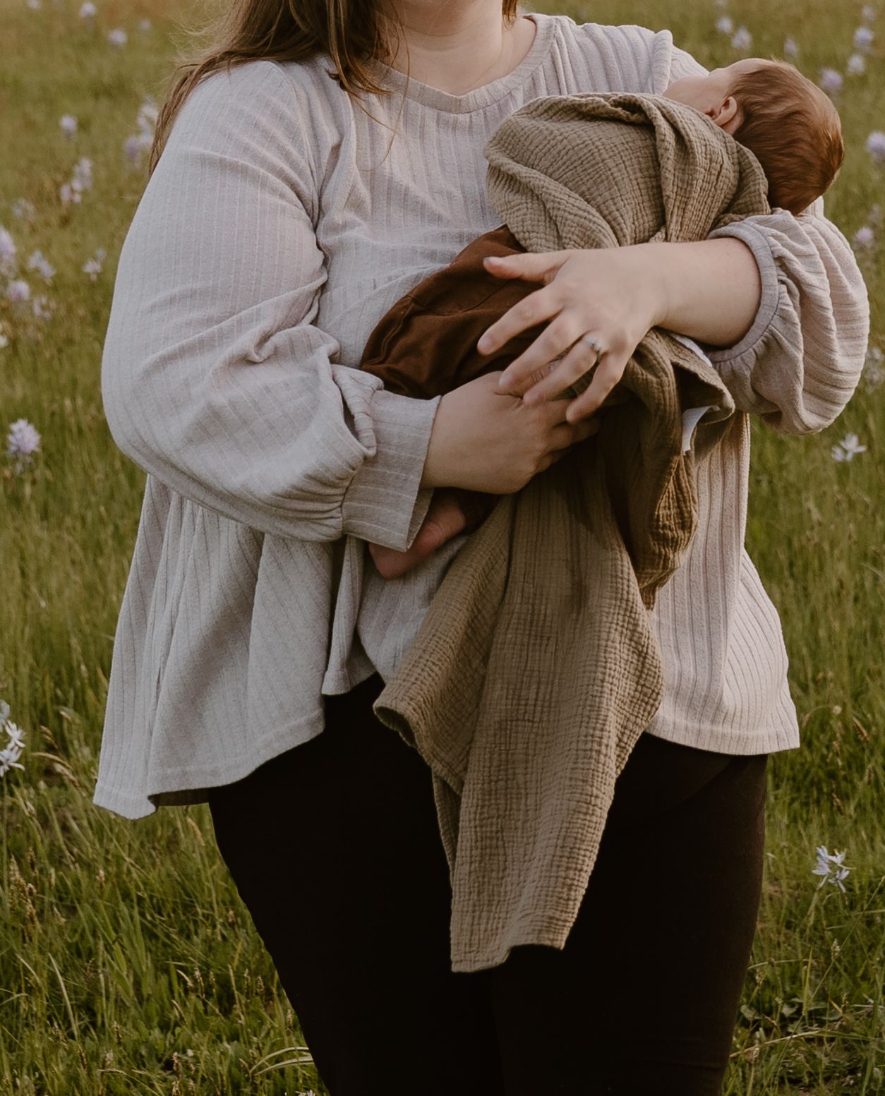 Mother holding newborn baby wrapped in tan muslin blanket while standing in a green wildflower field.