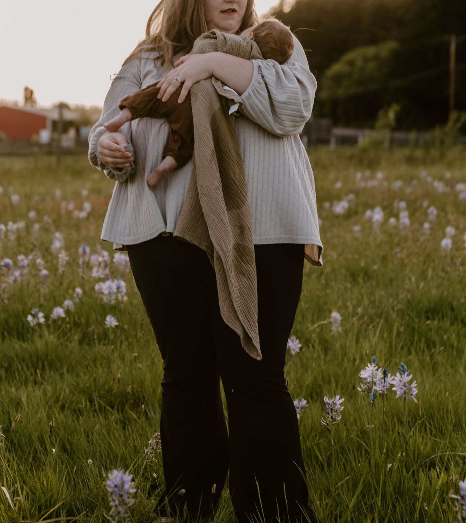 Woman holding two newborn babies in a wildflower field at golden hour, wearing a white ribbed sweater and black pants.
